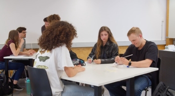 Im Vordergrund befinden sich drei Schüler:innen um einen Gruppentisch. In Hintergrund befindet sich eine andere Schülergruppe mit vier Personen an einem Tisch. Zwei davon stehen und die anderen sitzen.