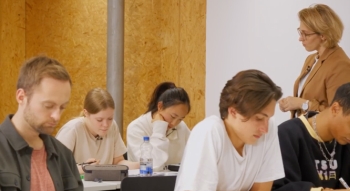 Auf dem Bild sind Schüler:innen in einem Klassenraum zu sehen. Die Lehrerin lehnt mit verschränkten Armen an der Wand und schaut in Richtung der Schüler:innen auf der letzten Bank hinten rechts.