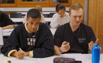 Auf dem Bild sind zwei Schüler zu sehen. Der linke Schüler hat einen Stift in der Hand und schaut in den Raum. Der rechte Schüler macht eine Geste.