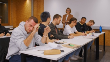 Auf dem Bild sind Schüler in einem Klassenraum zu sehe. Die Lehrerin steht seitlich des Klassenraums mit Blick Richtung Klasse. 