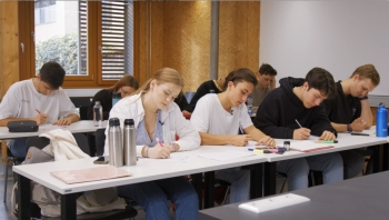 Auf dem Bild sind Schüler in einem Klassenraum zu sehen, die mit Stiften auf ein Blatt schreiben.