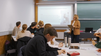 Klassenzimmers mit einer an die Rückwand projizierten Präsentation. Der Klasse zugewandt, steht davor eine Lehrerin in einem braunen Blazer. 