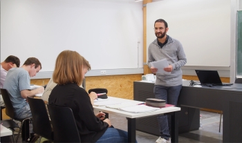 Lehrer der Klasse zugewandt vor einer weißen Tafel. Die Schüler:innen auf dem Bild schauen in Richtung des Lehrers.