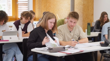 Zwei Schüler:innen arbeiten einander zugewandt. Im Hintergrund sind weitere Schüler:innen zu sehen, die arbeiten.