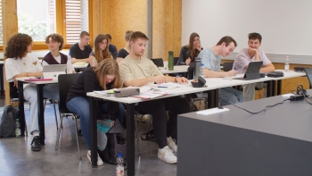 Foto eines Klassenzimmers. Eine Schülerin in der ersten Sitzreihe greift in ihren Rucksack.