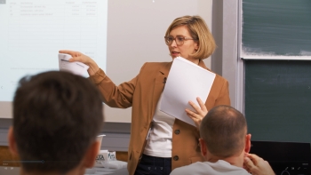 Lehrerin mit braunem Blazer steht vor einem projiziertem Bild, auf das sie zeigt. In ihrer linken Hand hält sie Papier.