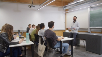 Lehrer mit grauem Pullover steht vor einer weißen Tafel und ist der Klasse zugewandt. Die Schüler:innen schauen in seine Richtung.