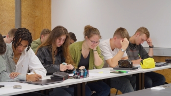 Auf dem Foto ist eine Klasse von Schüler:innen zu sehen, die in einer Gruppe an einem Tisch zusammensitzen.