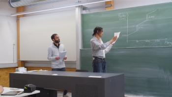 Auf dem Foto ist zu sehen, wie ein Schüler an die Tafel schreibt. Der Lehrer steht hinter dem Schüler und schaut auf die Tafel.