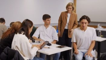 Lehrerin mit braunem Blazer steht an einem Arbeitstisch der Schüler:innen. Ein Schüler dreht sich weg von den anderen weg.Schüler:innen.