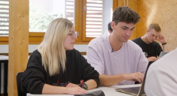 Auf dem Foto sind eine Schülerin und ein Schüler zu sehen. Die Schülerin schaut auf den Laptop ihres Nachbarn.