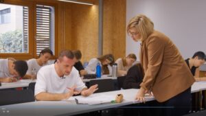 Lehrerin mit braunem Blazer steht vor einem Tisch und ist einem Schüler zugewandt. Im Hintergrund des Fotos arbeiten weitere Schüler:innen.